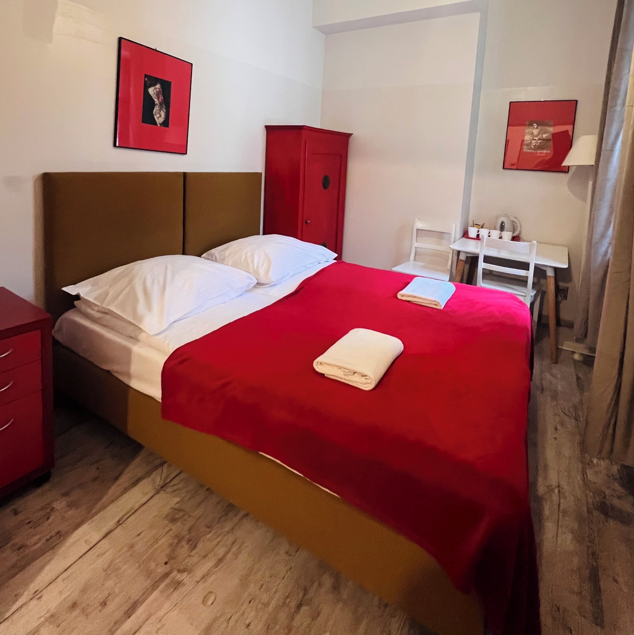 Ein ordentlich bezogenes Bett mit weißer und roter Bettwäsche, ein roter Kleiderschrank und ein kleiner Tisch mit einem Stuhl.