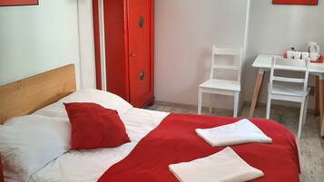 Ett hotellrum med en säng, en röd garderob, ett skrivbord med en stol och ett bord med en vattenkokare och koppar.