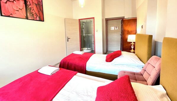 A hotel room with two beds, each with red bedding and pillows, a wooden headboard, a bedside lamp, and a shower area visible through a glass door. - Oki Doki OLD TOWN (Warsaw)