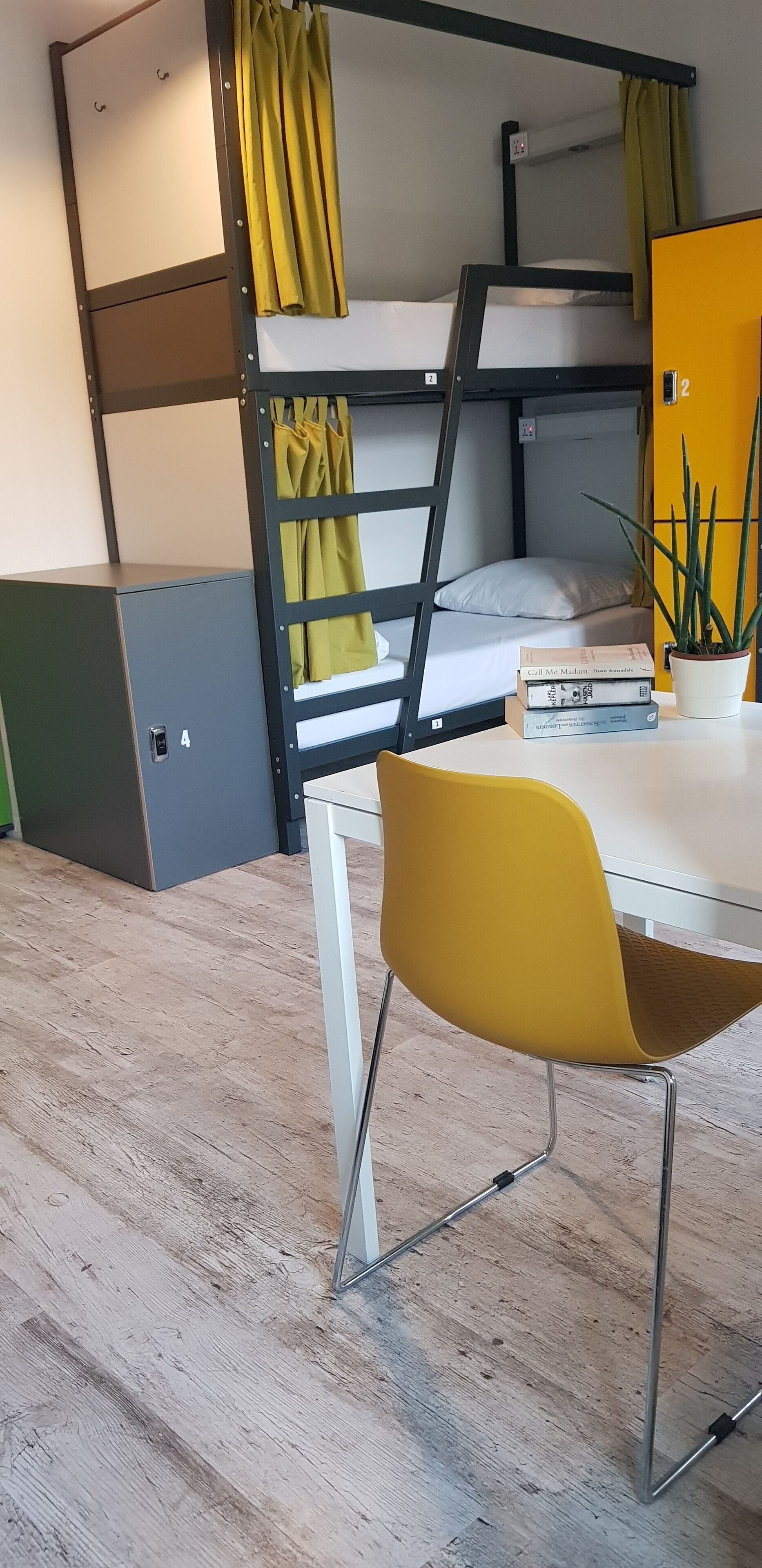 Een stapelbed met een geel gordijn, een kleine tafel met een plant en een gele stoel in een kamer met een houten vloer.