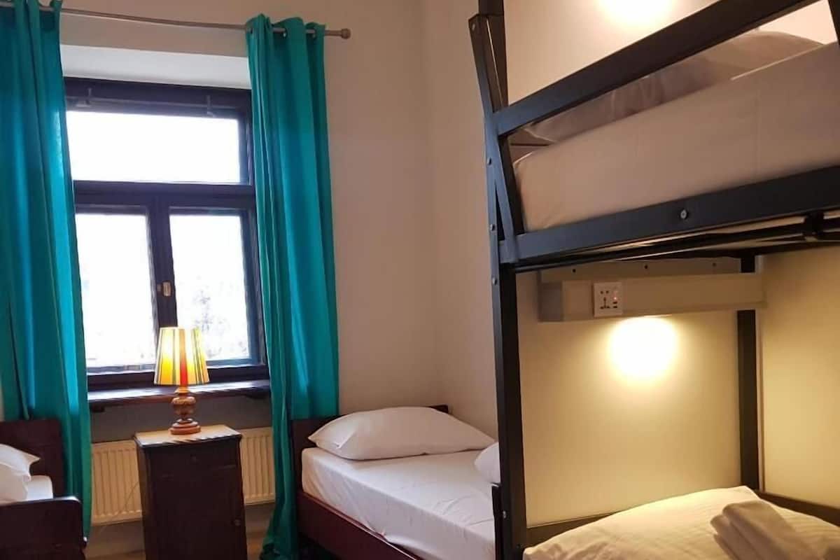 A bunk bed room with a window, curtains, a bedside table with a lamp, and a radiator.