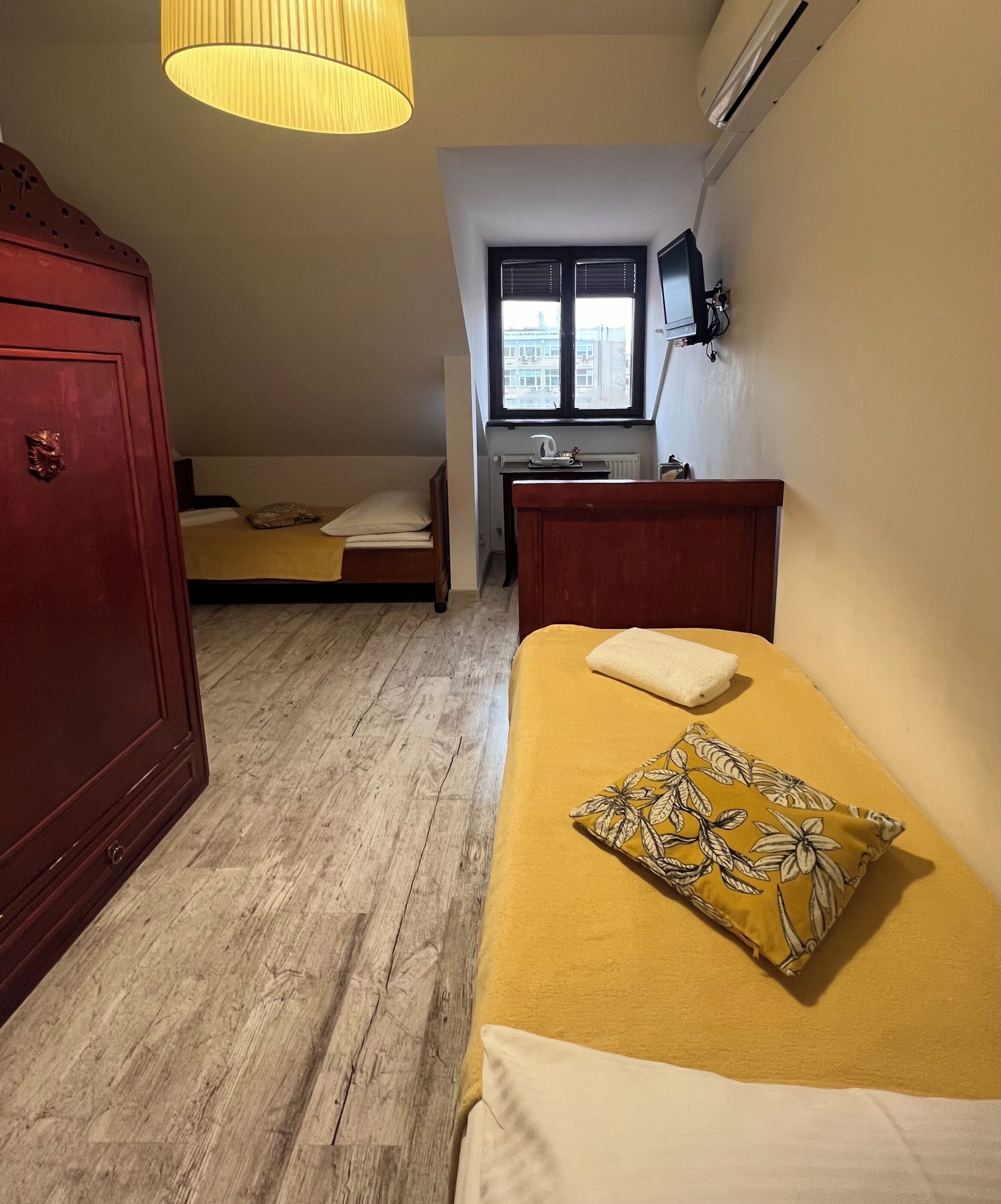 A compact hotel room with a wooden floor, a bed with a yellow blanket and a floral pillow, a wooden wardrobe, a desk with a chair, a window with blinds, and an air conditioning unit.