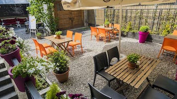 Un patio al aire libre con mesas y sillas, rodeado de plantas en macetas y un paraguas grande.