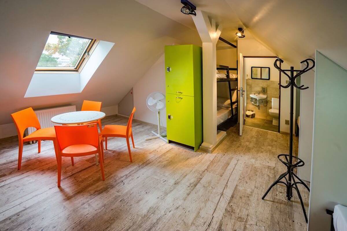 A room with a sloped ceiling, a skylight, a green wardrobe, a round table with orange chairs, and a coat rack.