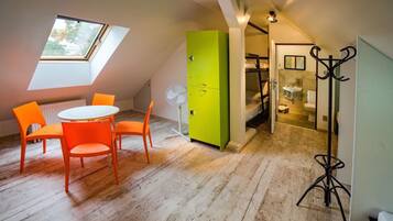 A room with a sloped ceiling, a skylight, a green wardrobe, a round table with orange chairs, and a coat rack.