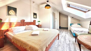 Une chambre à coucher avec un grand lit, un plus petit lit, un bureau et une chaise. Il y a une fenêtre qui donne sur le ciel.