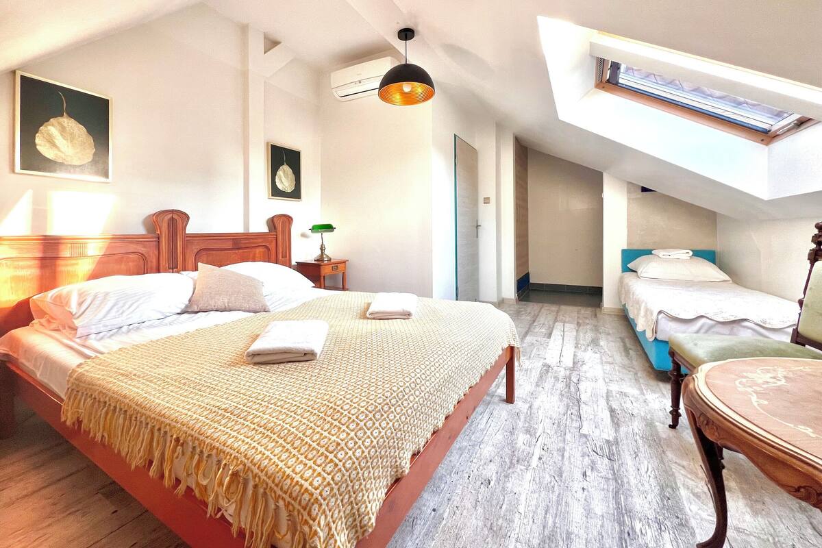 A bedroom with a large bed, a smaller bed, a desk, and a chair. There is a window with a view of the sky.