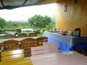 An outdoor dining area with wooden benches and tables, a covered section with a table set for a meal, and a view of greenery and mountains in the background. - Baanrai Areeya (Suan Phueng)