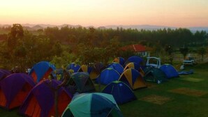 A campsite with multiple colorful tents arranged on a grassy field, surrounded by trees and a distant mountain range. - Baanrai Areeya (Suan Phueng)