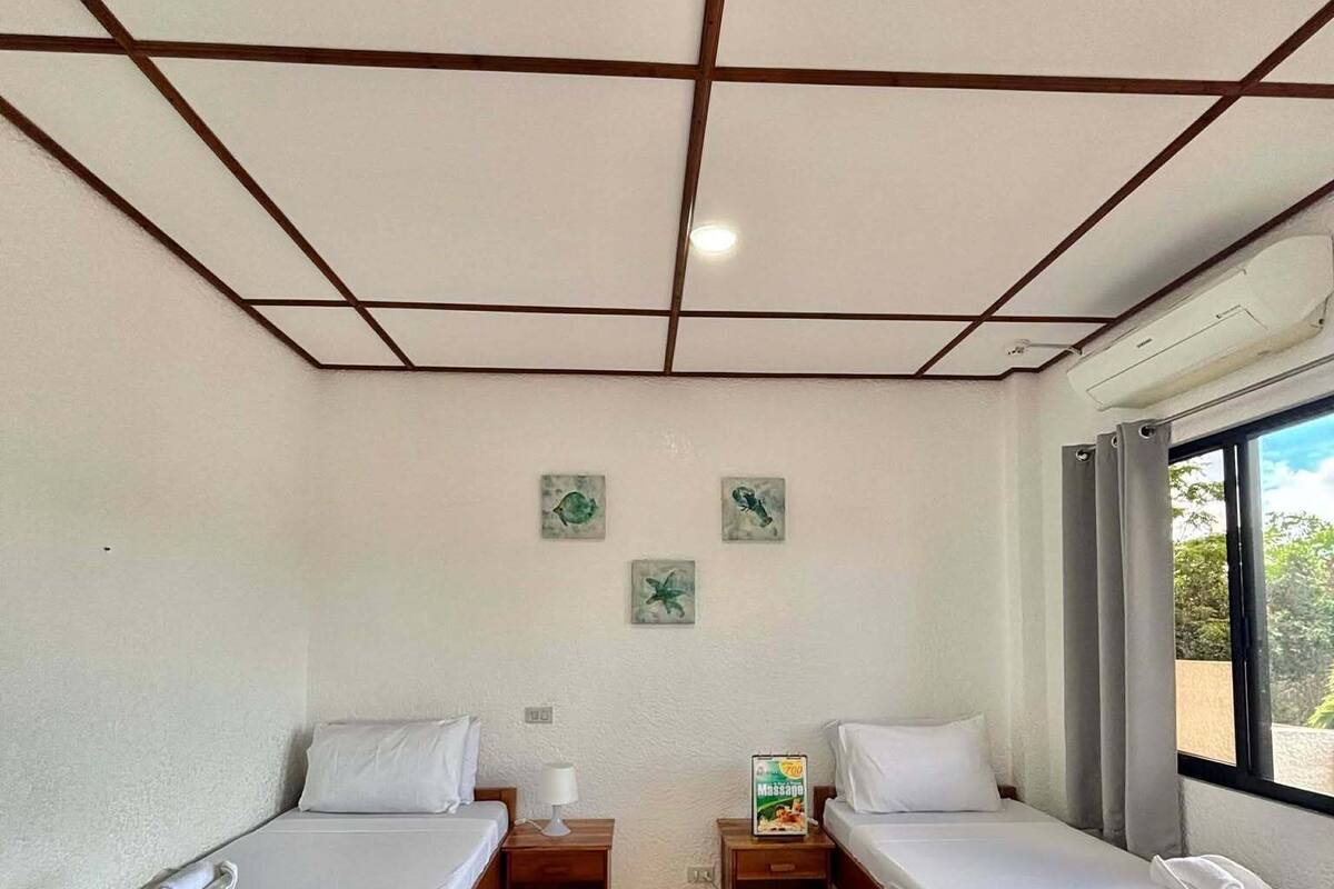 Ein Hotelzimmer mit zwei Betten, einem kleinen Tisch, einem Nachttisch, einem Fenster mit Vorhängen und einer Decke mit Rastermuster.