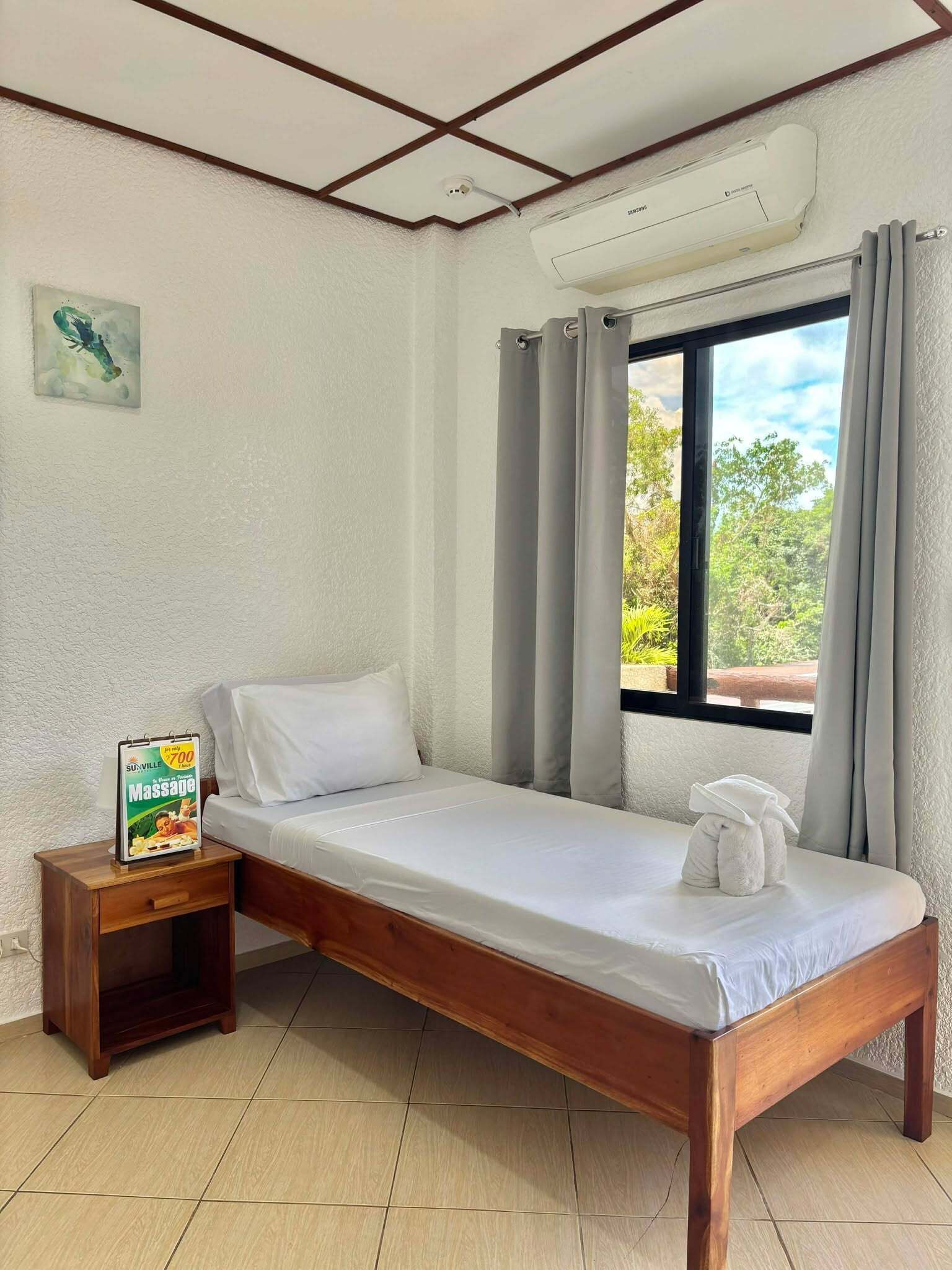 A single bed with a wooden frame, a nightstand with a sign, a window with curtains, and a ceiling with a recessed light fixture.
