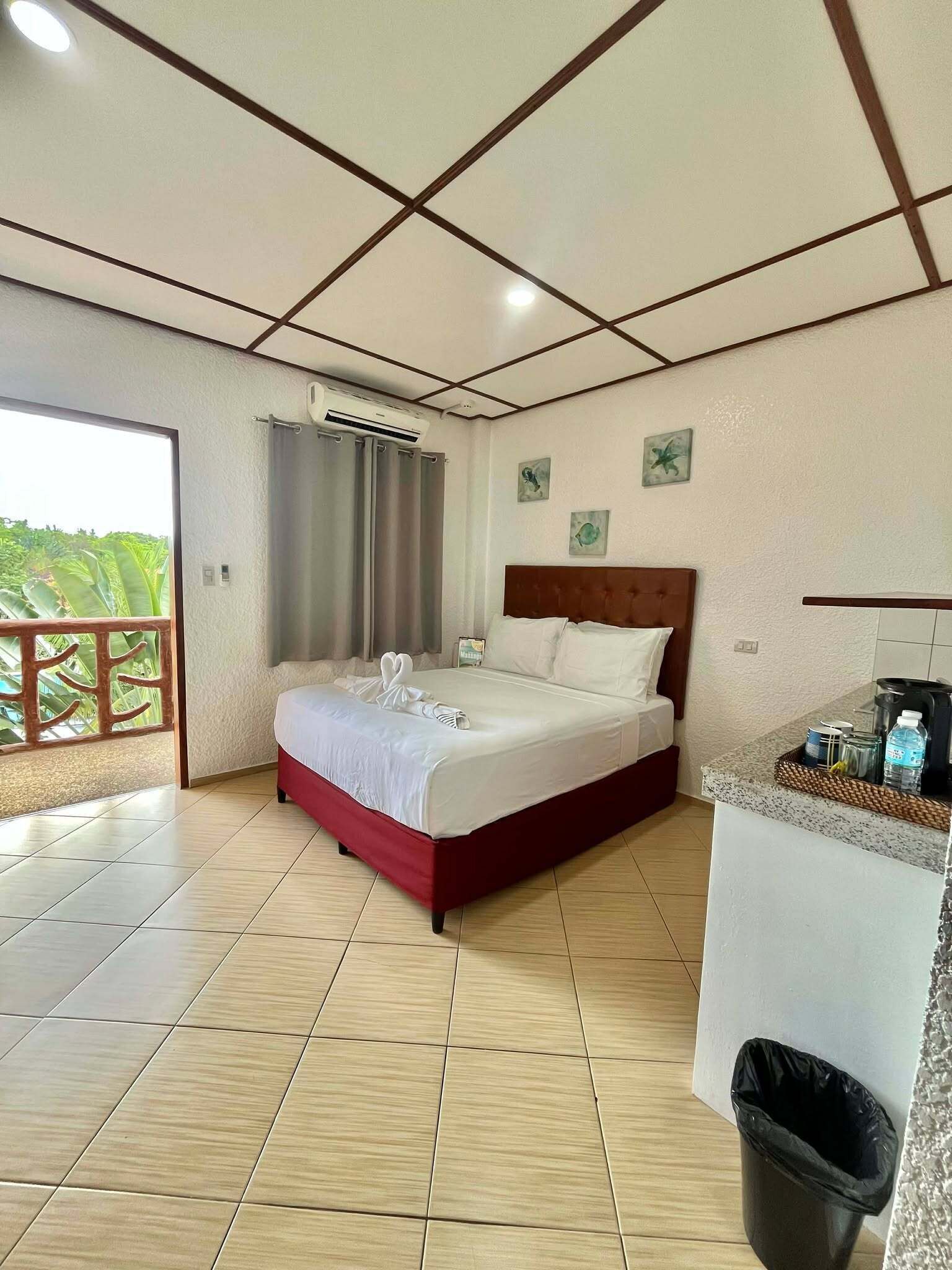 A hotel room with a bed, a small kitchenette, a trash bin, and a balcony with a view.