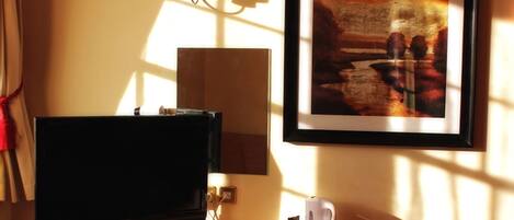 Habitación de hotel con televisión, un cuadro en la pared, una lámpara y una mesita con control remoto, una taza y una caja de pañuelos.