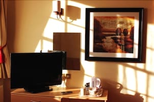 Quarto de hotel com televisão, um quadro na parede, uma luminária e uma mesinha com controle remoto, uma xícara e um estojo de lenços.