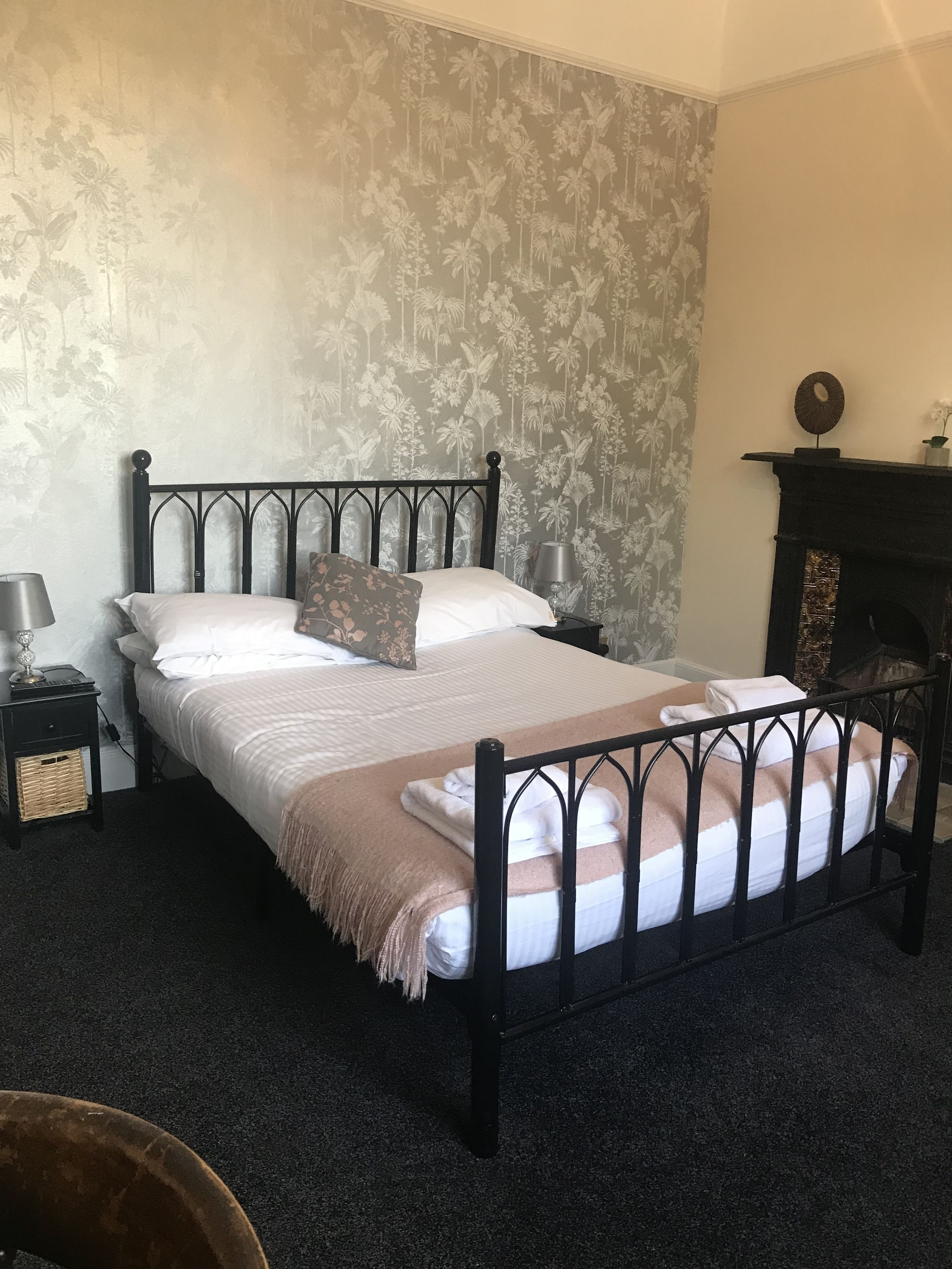 A neatly made bed with a headboard, a bedside table with a lamp, and a fireplace in the corner.