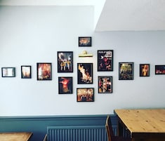 Paredes com várias imagens emolduradas, um candeeiro e uma mesa de madeira.