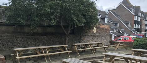 Mesas de picnic de madera al aire libre, dispuestas sobre una zona pavimentada con un muro de piedra y un árbol al fondo.