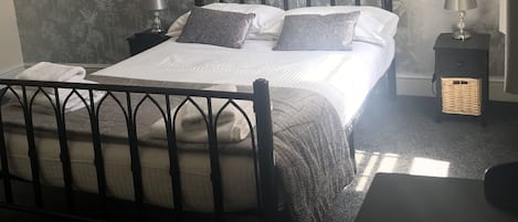 A neatly made bed with a black metal frame, white bedding, and two pillows. There is a small bedside table with a lamp and a decorative item. The room has a floral wallpaper and a dark carpet.