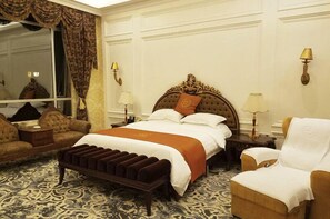 Habitación de hotel con una cama grande, zona de estar con sofá y sillón, un banco y dos mesitas de noche con lámparas.