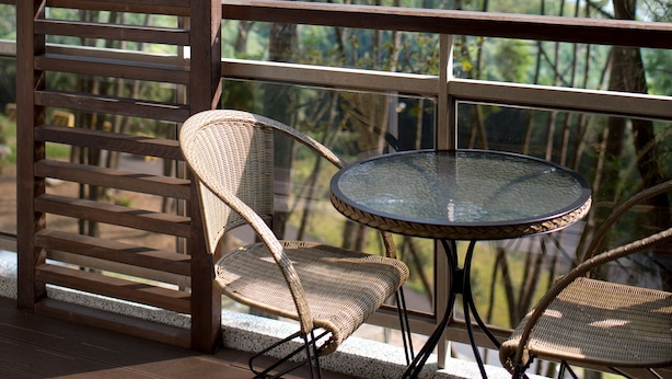Een balkon met een glazen tafel en rieten stoelen, met uitzicht op een bebost gebied.