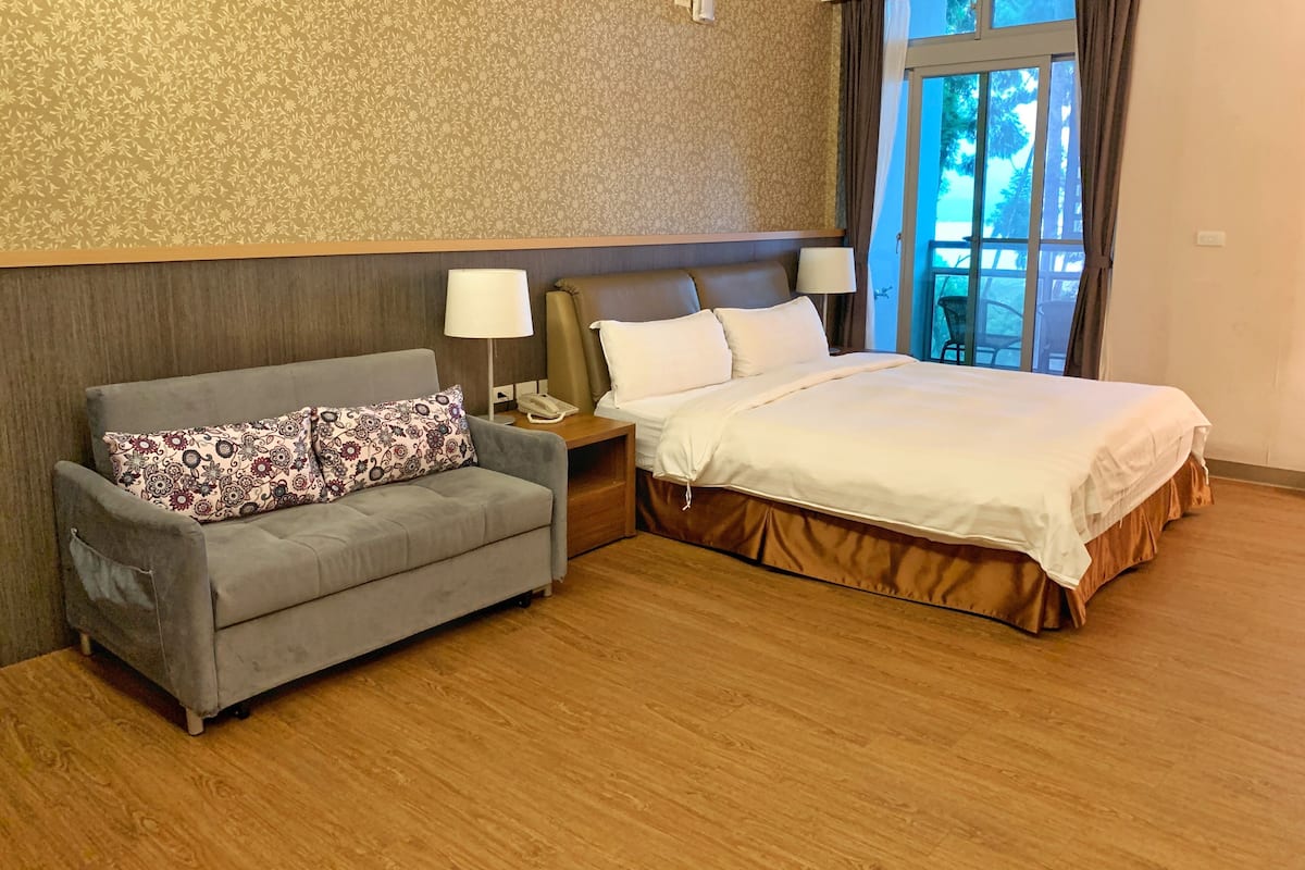 A hotel room with a bed, a gray sofa, two bedside lamps, a telephone, and a window with curtains.