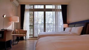 Een hotelkamer met twee bedden, een bureau met stoel, een lamp en een groot raam met jaloezieën.