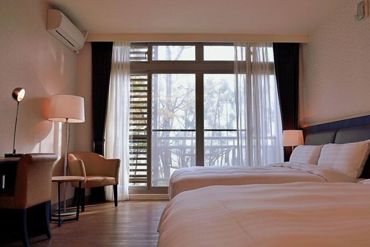 A hotel room with two beds, a desk with a chair, a lamp, and a large window with blinds.