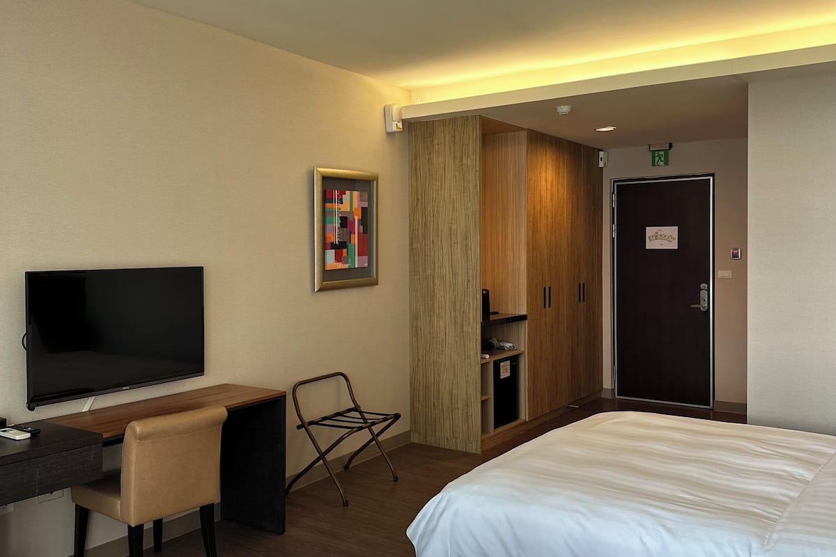 A hotel room with two beds, a desk with a chair, a flat-screen TV, a wooden wardrobe, and a door with an exit sign.