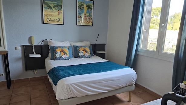 Quarto com uma cama, duas pinturas penduradas na parede, uma mesa de cabeceira com abajur e uma janela com cortinas.