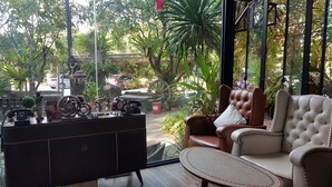 A room with a leather sofa, a wooden cabinet with various items, and a view of a garden with trees and a stone structure. - Rayonghouse Resort (Rayong)