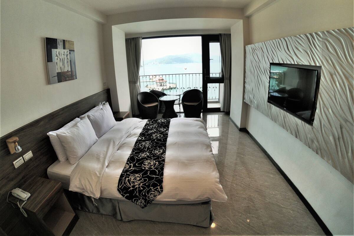 Une chambre d’hôtel avec un lit, un téléviseur fixé au mur, un balcon avec vue et une petite table avec des chaises.