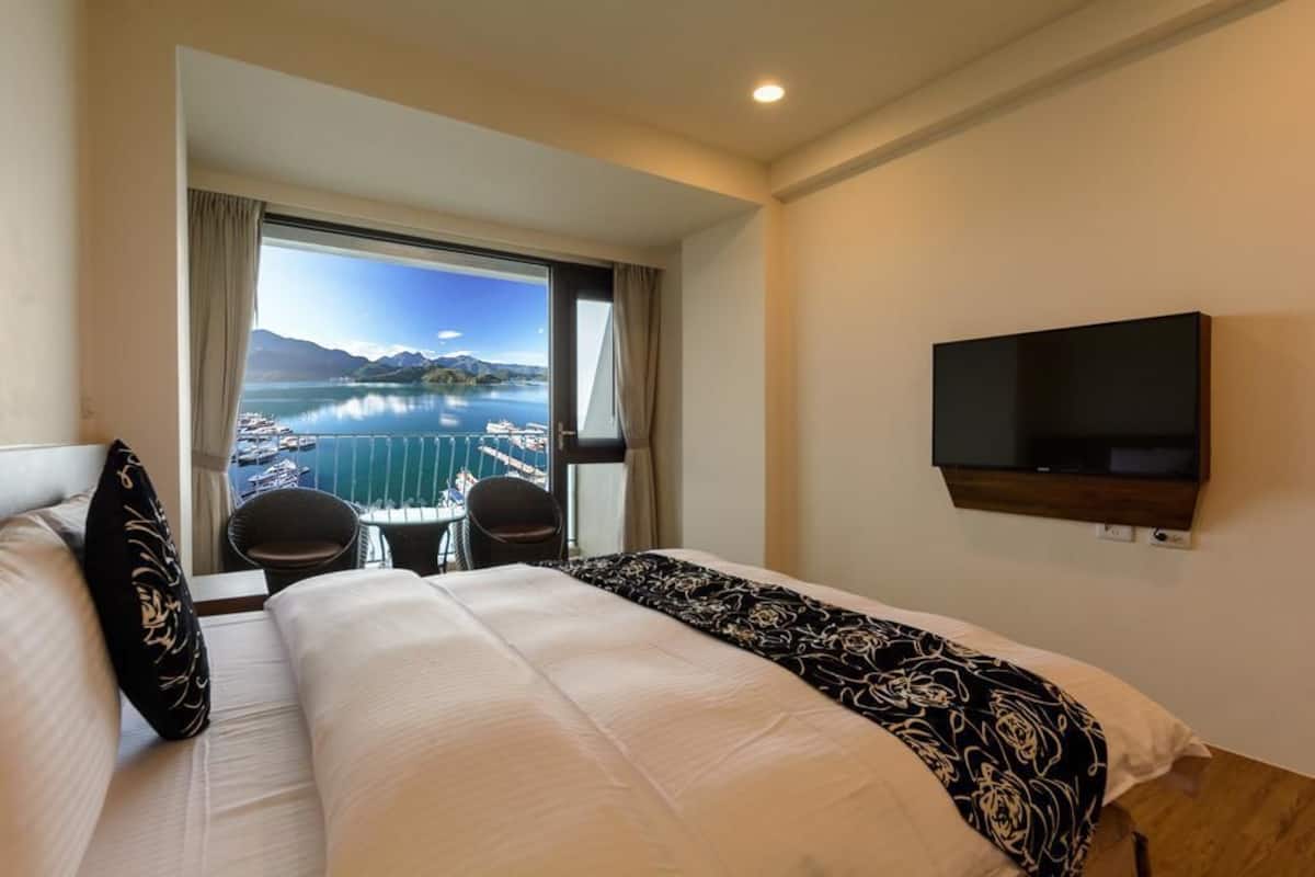 Une chambre d’hôtel avec un grand lit, un téléviseur fixé au mur et une vue sur un lac et des montagnes, visible par la fenêtre.