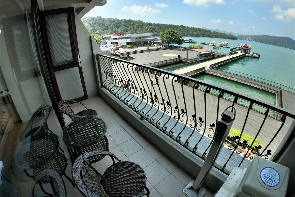 Un balcon avec des fauteuils en osier et une table, donnant sur un front de mer où l’on peut voir des bateaux et un quai.