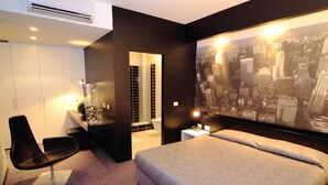 A modern hotel room with a large bed, a desk with a chair, and a cityscape mural on the wall. - Design Suite Tirano (Tirano)