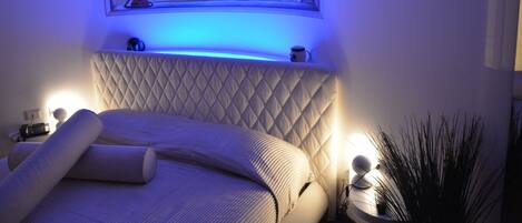 Un dormitorio con cama, mesita de noche, una obra de arte en la pared que representa un velero y una mesilla con lámpara.