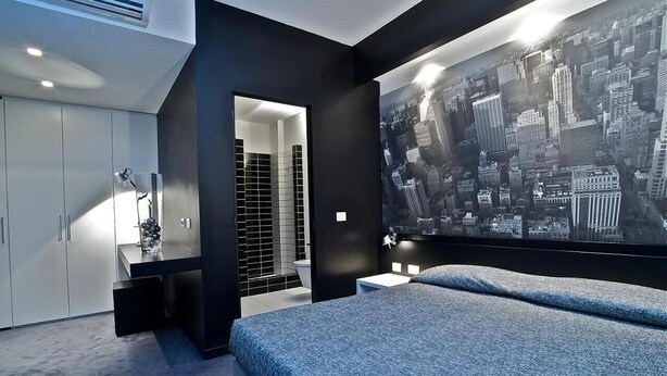 A modern bedroom with a large bed, a cityscape mural, a walk-in closet, and a bathroom with black tiles. - Design Suite Tirano (Tirano)