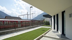 A modern building with a covered outdoor area, a train on the adjacent track, and a mountain in the background.