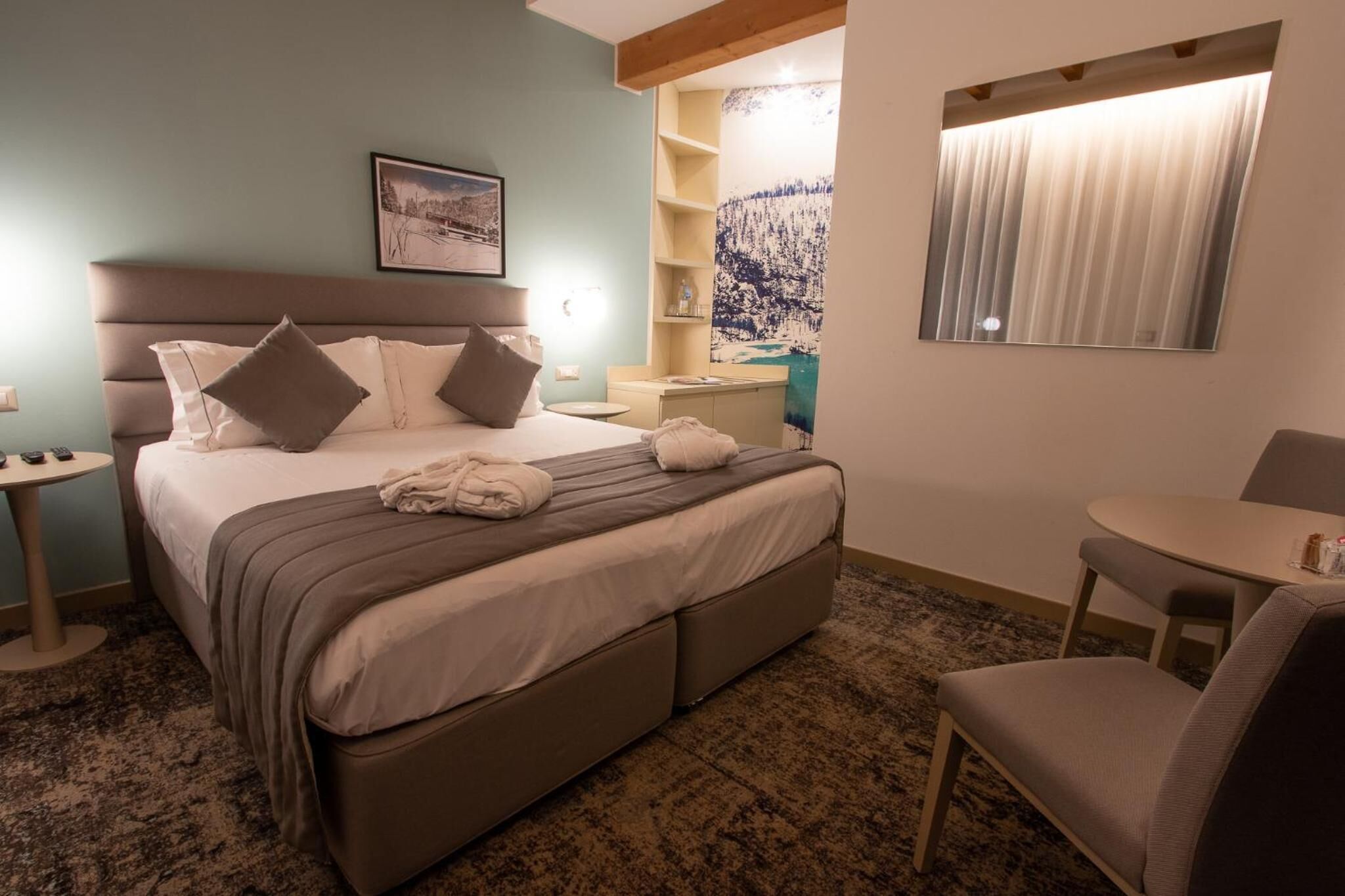 A hotel room with a bed, a chair, a small table, a nightstand, a shelf with decorative items, and a painting on the wall.