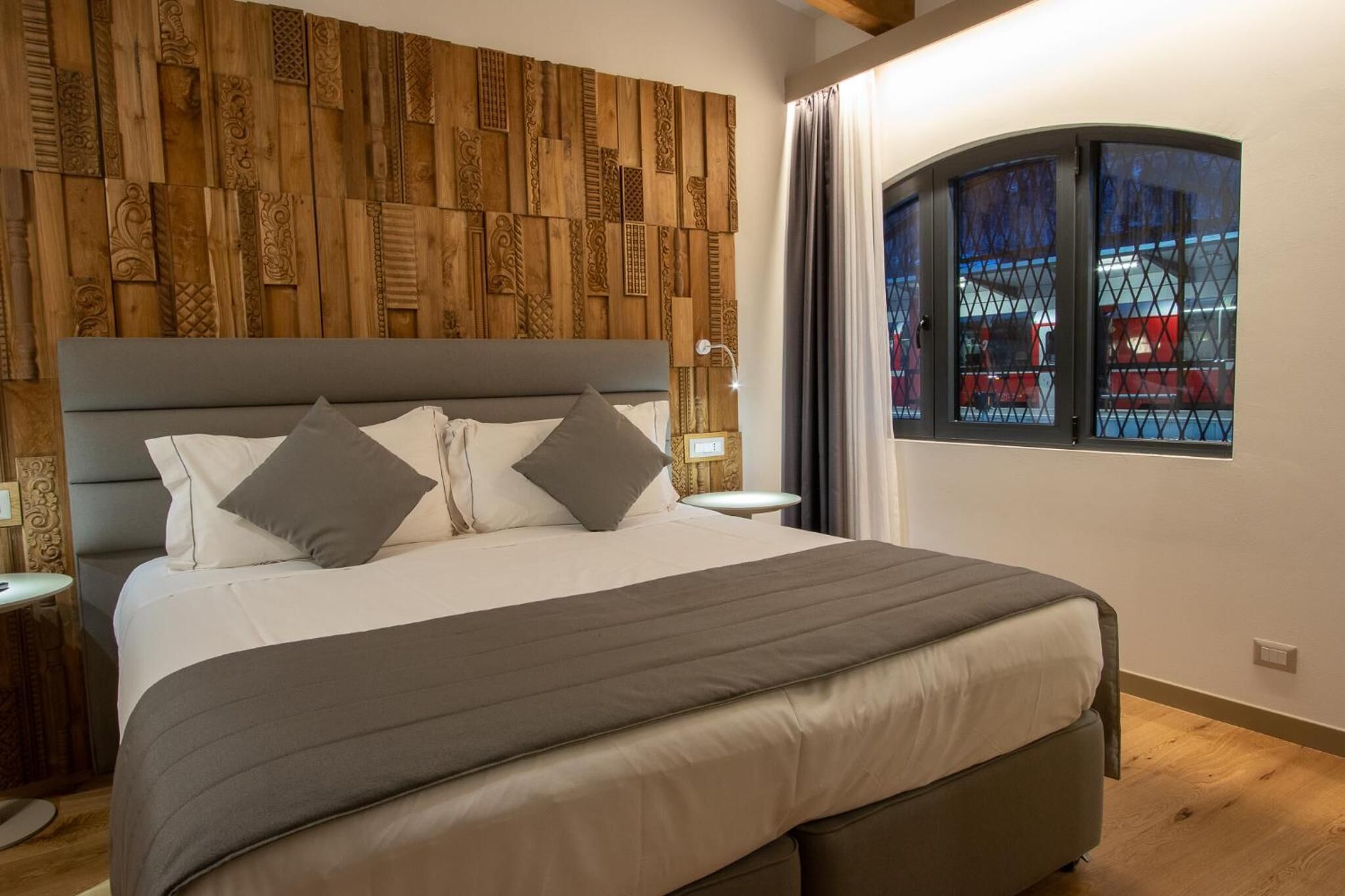 A neatly made bed with a grey headboard and pillows, a wooden wall panel, a bedside table with a lamp, and a window with a view of a red vehicle outside.