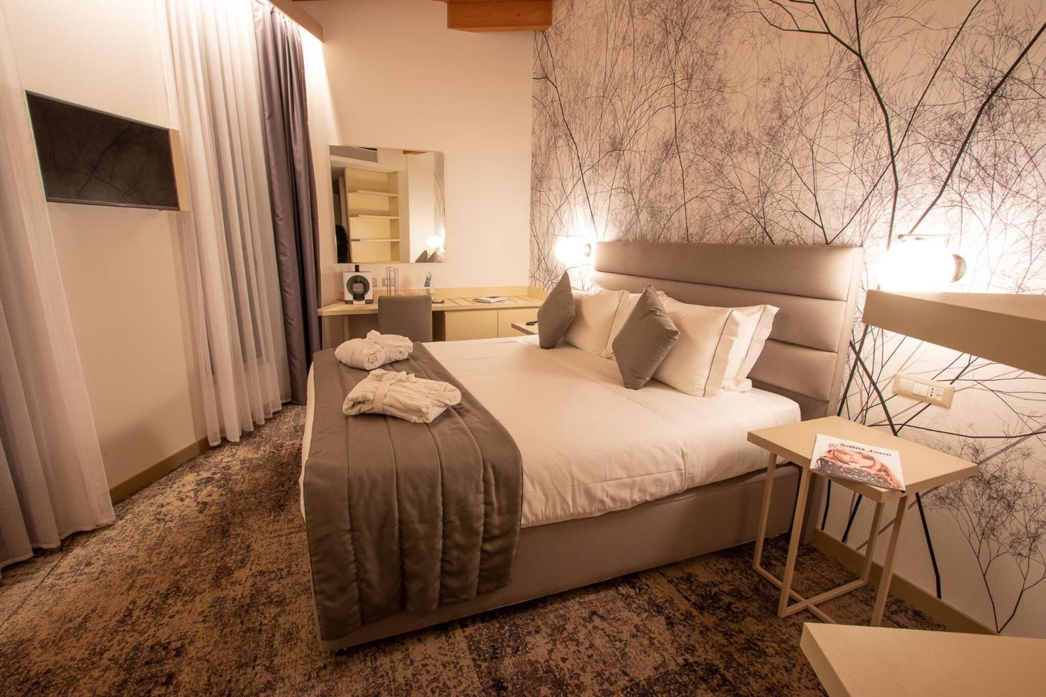 Habitación de hotel con una cama grande, una mesita de noche, un escritorio con una silla, un espejo y una ventana con cortinas.