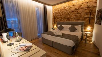 Une chambre d’hôtel moderne avec un mur en bois à motifs en chevrons, un grand lit avec une literie grise, une table de chevet avec une lampe et une vue sur la ville à travers des rideaux transparents.