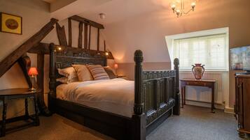 A bedroom with a wooden four-poster bed, a nightstand with a lamp, a small table with a vase, and a window with blinds.