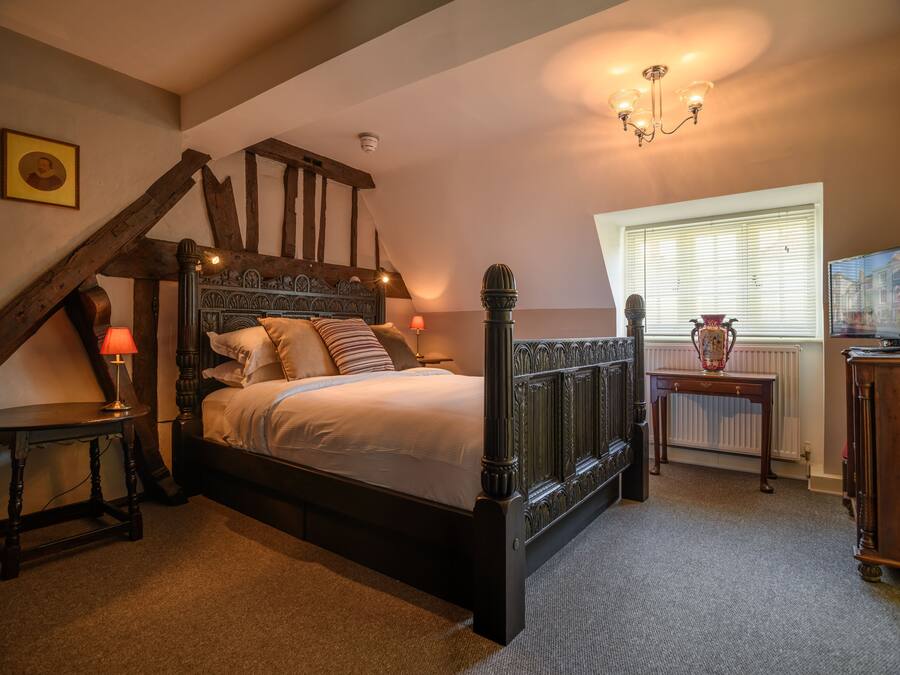A bedroom with a wooden four-poster bed, a nightstand with a lamp, a small table with a vase, and a window with blinds.