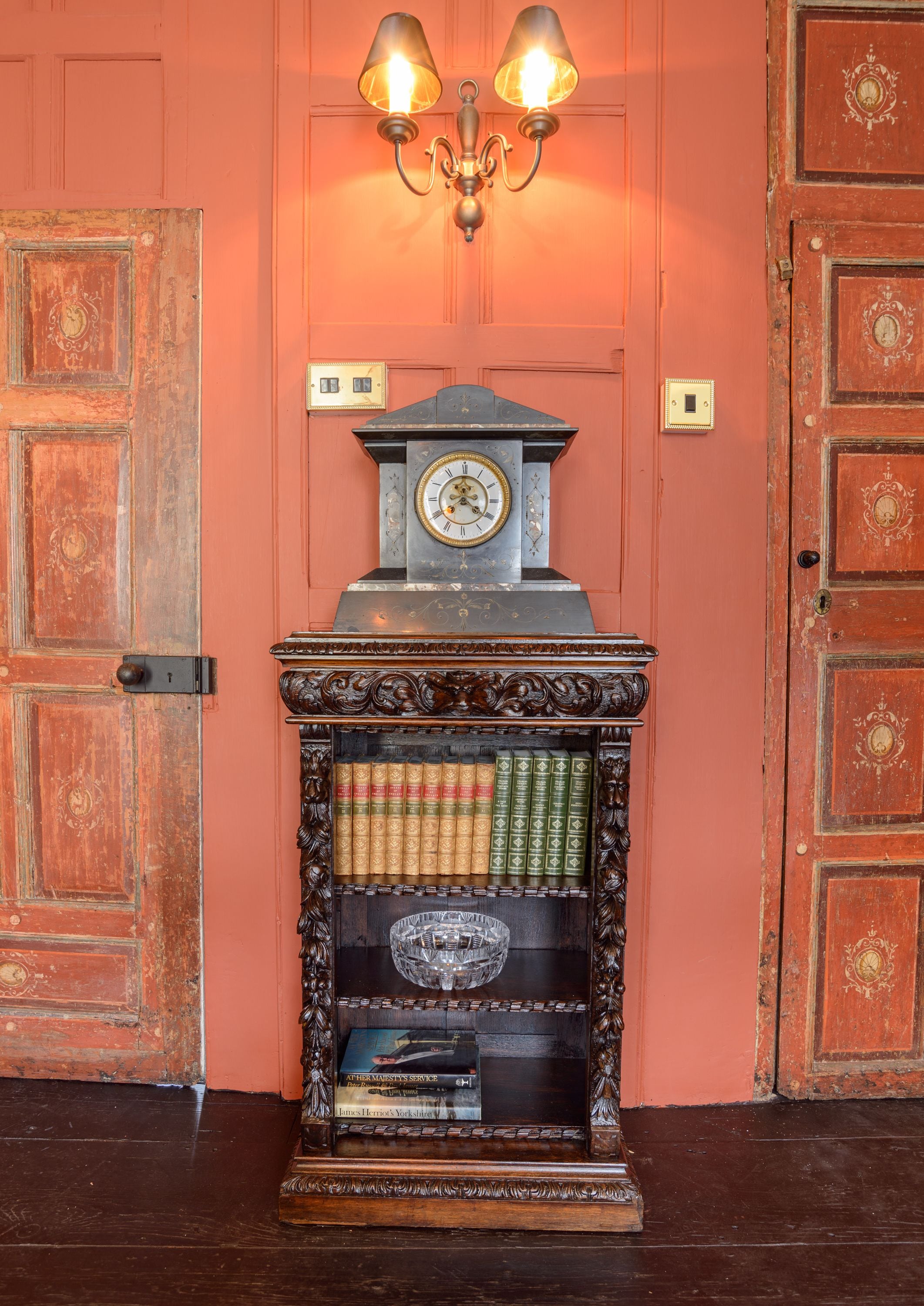 A vintage wooden bookshelf with a clock and books, against a peach-colored wall with two wooden doors.