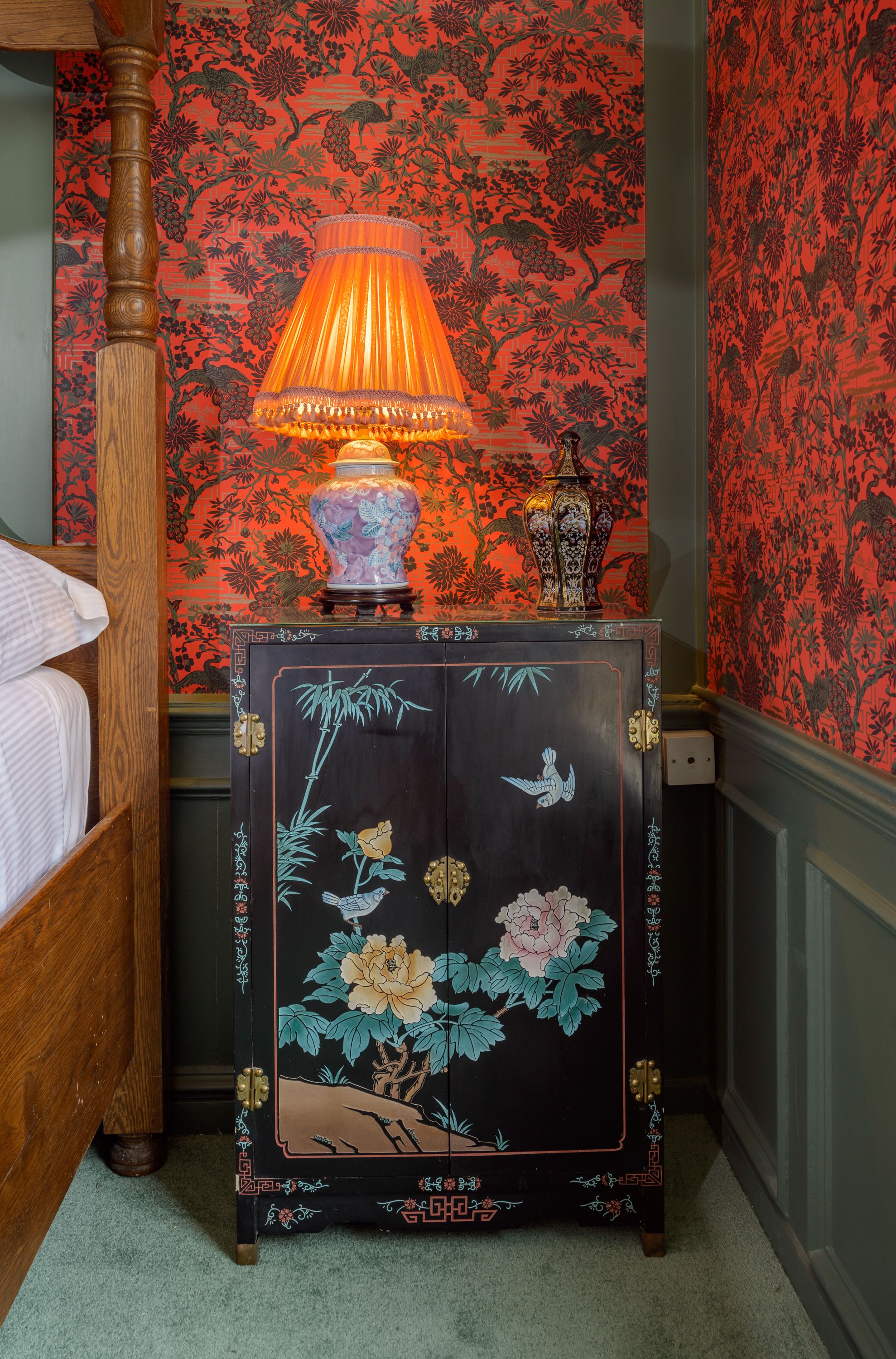A bedroom with a wooden four-poster bed, a nightstand with a lamp, a decorative vase, and a cabinet with floral artwork.