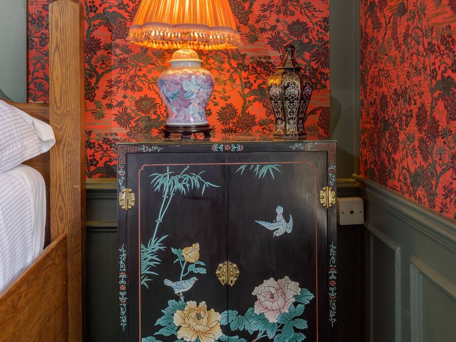 A bedroom with a wooden four-poster bed, a nightstand with a lamp, a decorative vase, and a cabinet with floral artwork.