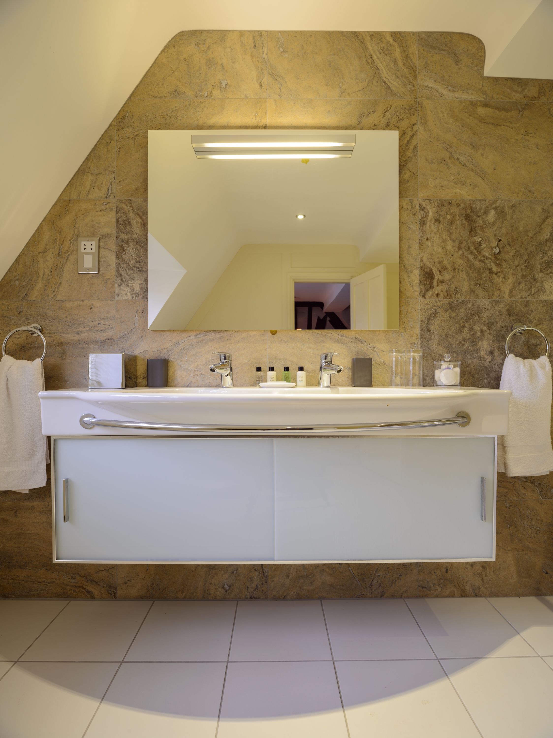A bathroom with a stone wall, a large mirror, a double sink with a towel ring, and a cabinet below.