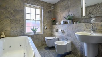 A bathroom with a large bathtub, a toilet, and a sink. There is a window with a view of the outside, and a shelf with toiletries.