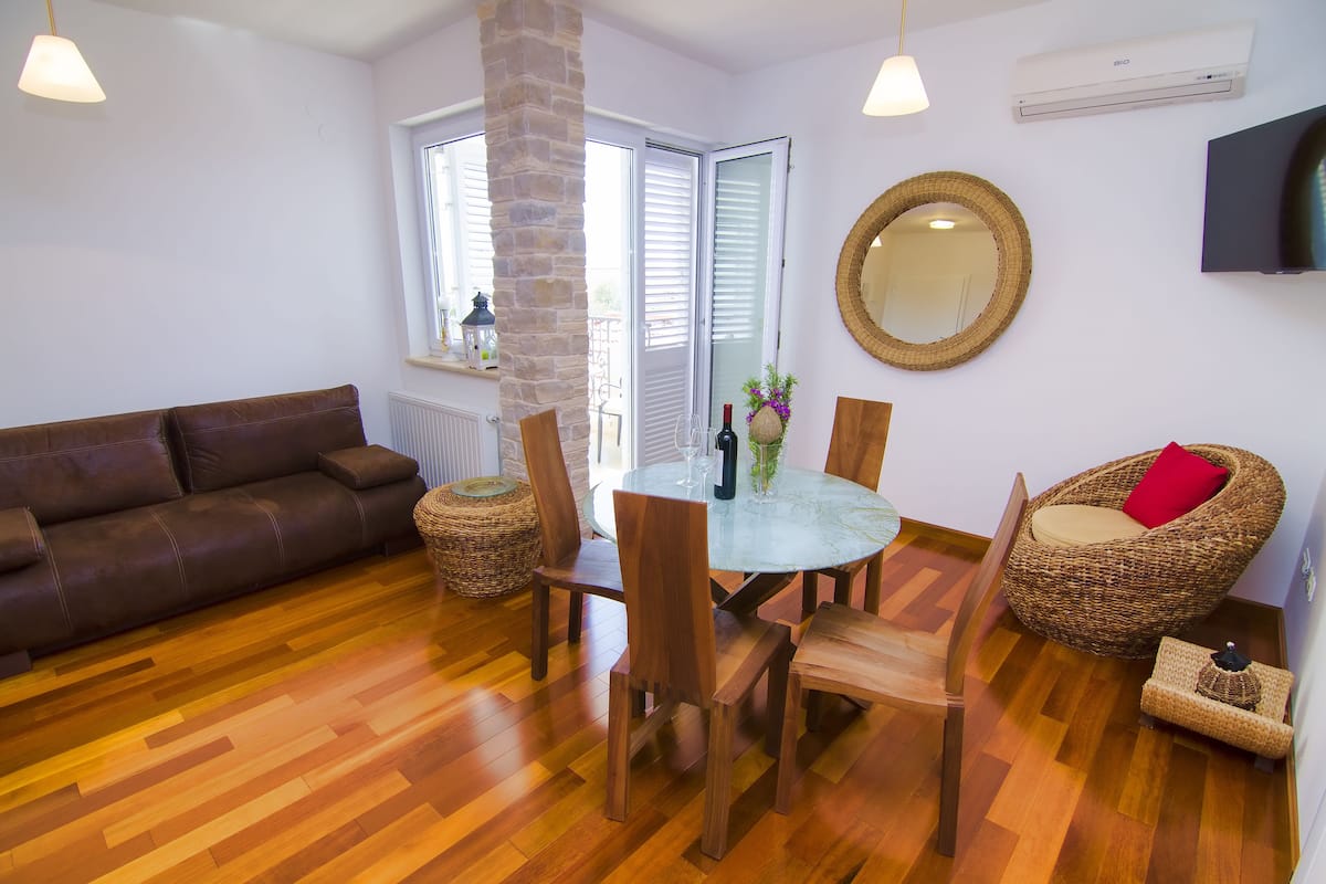 A cozy living area with a brown leather sofa, wooden dining table with chairs, wicker armchairs, and a large mirror on the wall.