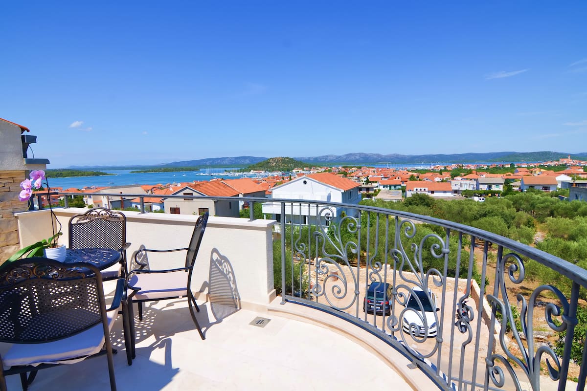 A balcony with a view of a coastal town, outdoor seating, and a railing.